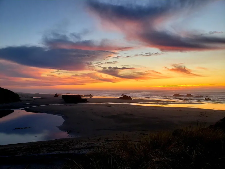 Bandon Beach at sunset with sea stacks scattered in the surf, dune grass in the foreground, and tidepool reflections holding the color of the sky