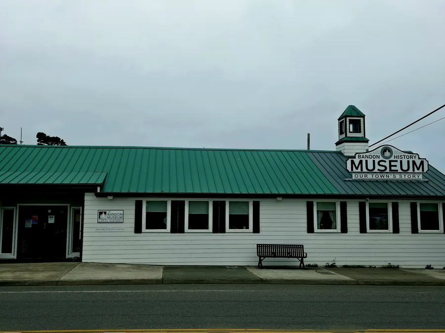 The Bandon History Museum, a long white clapboard building with a green metal roof and a small cupola, on the corner of Highway 101 and Fillmore Avenue under an overcast coastal sky