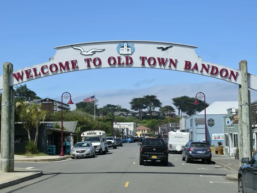 The Welcome to Old Town Bandon archway over First Street, with the cranberry-red lettering and lighthouse medallion overhead and the businesses of Old Town under it